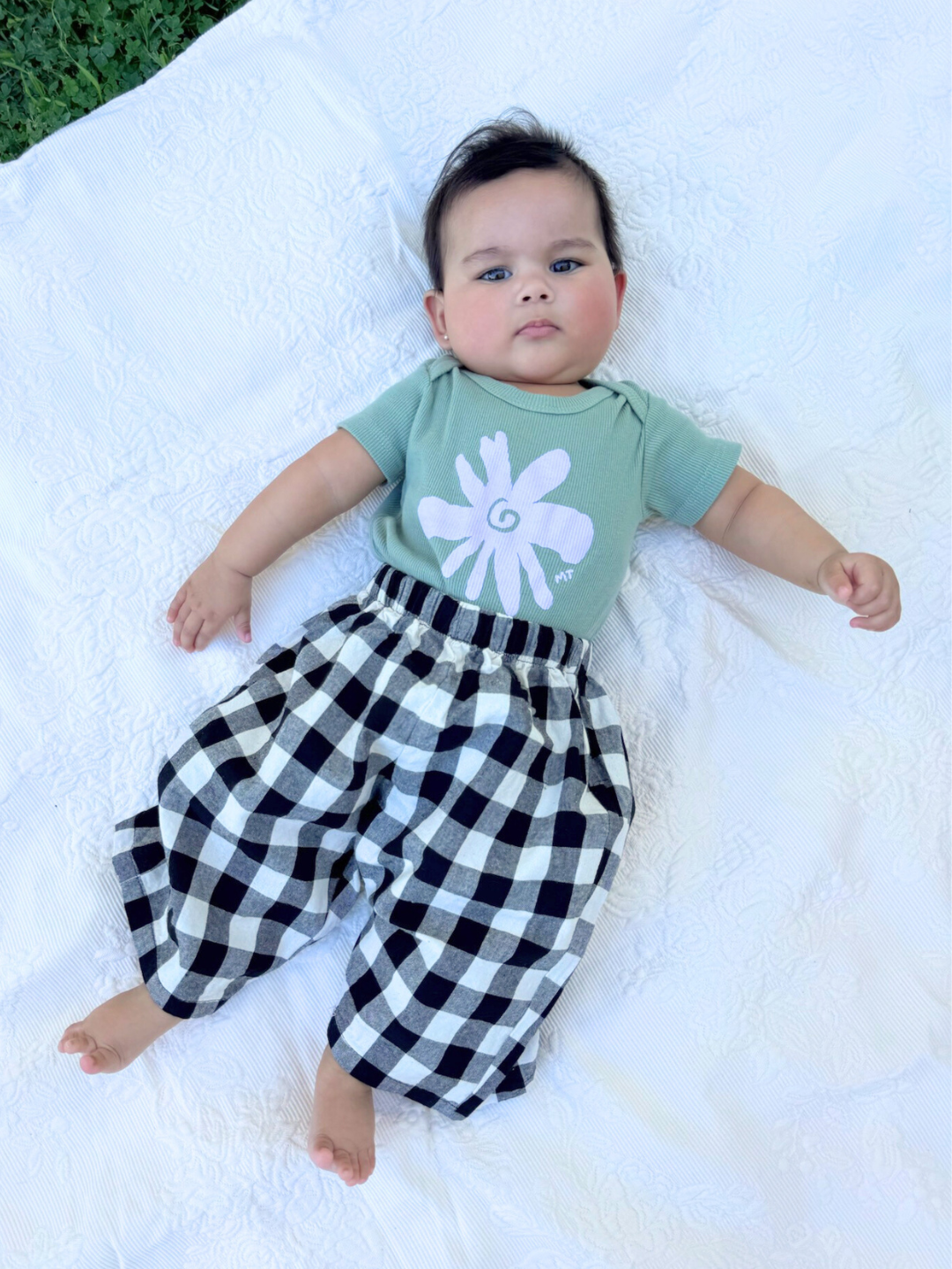 Green | A baby wearing the flower onesie, in sage green with a light purple flower on the front. She wears black and white gingham pants and lies on a white blanket.