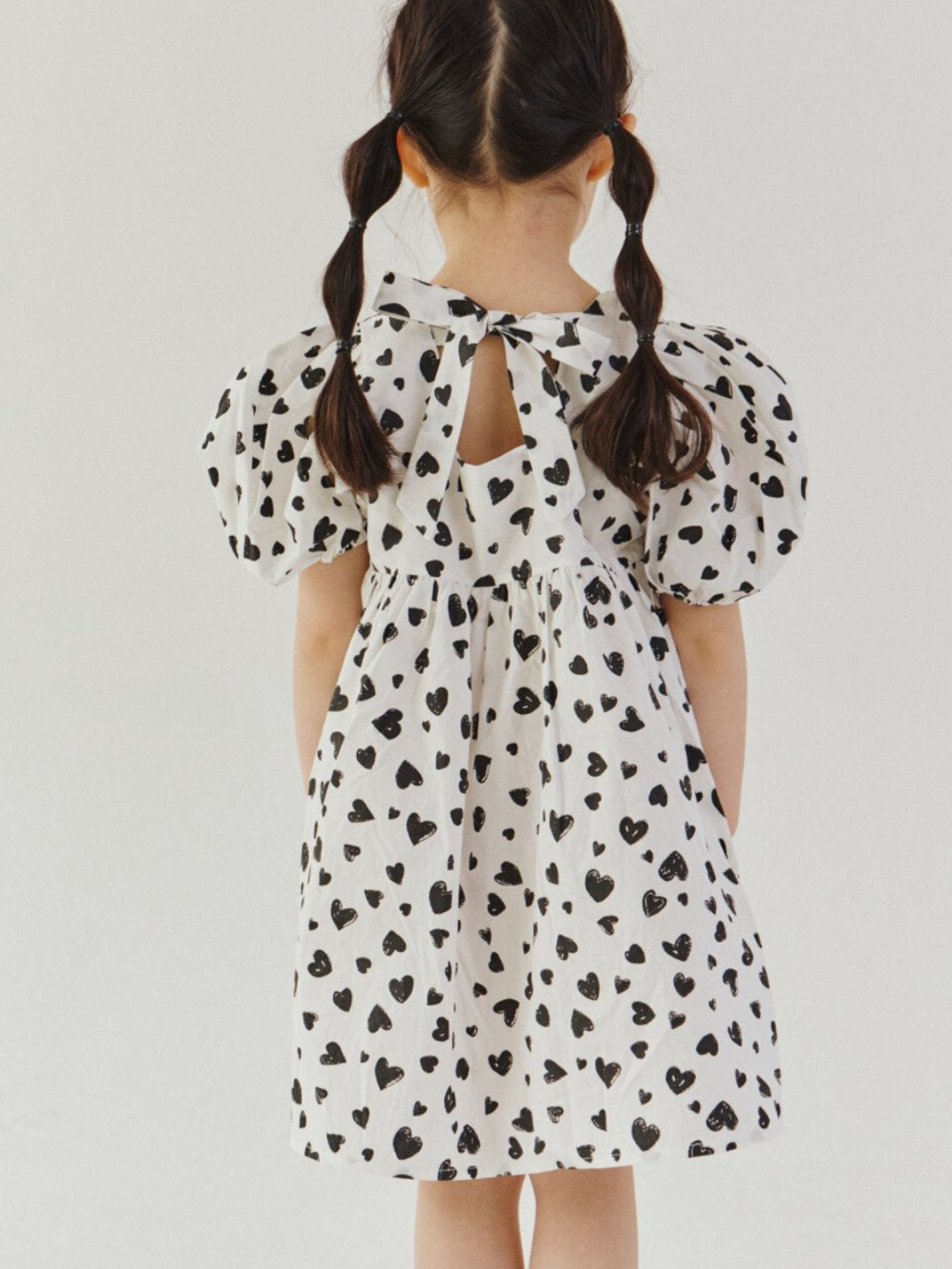 A child wearing a kids' puff-sleeved dress with black hearts on a white background, with bow tied at the neck, back view