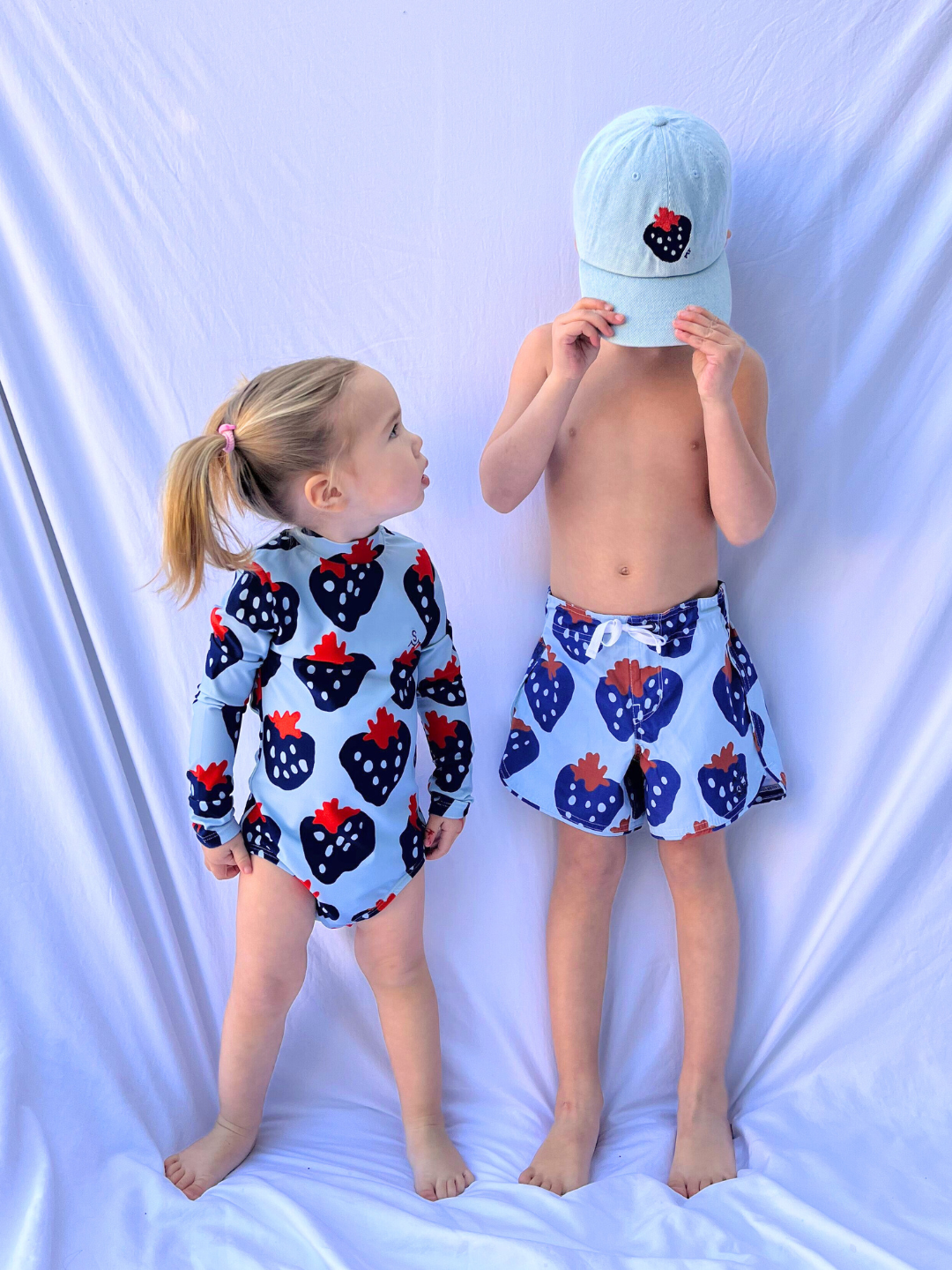 Two children in strawberry-patterned swimsuits stand against a white backdrop. The girl wears a long-sleeve, sustainably-made suit. The boy sports blue STRAWBERRY BOARDSHORTS and a cap, adjusting it over his face.