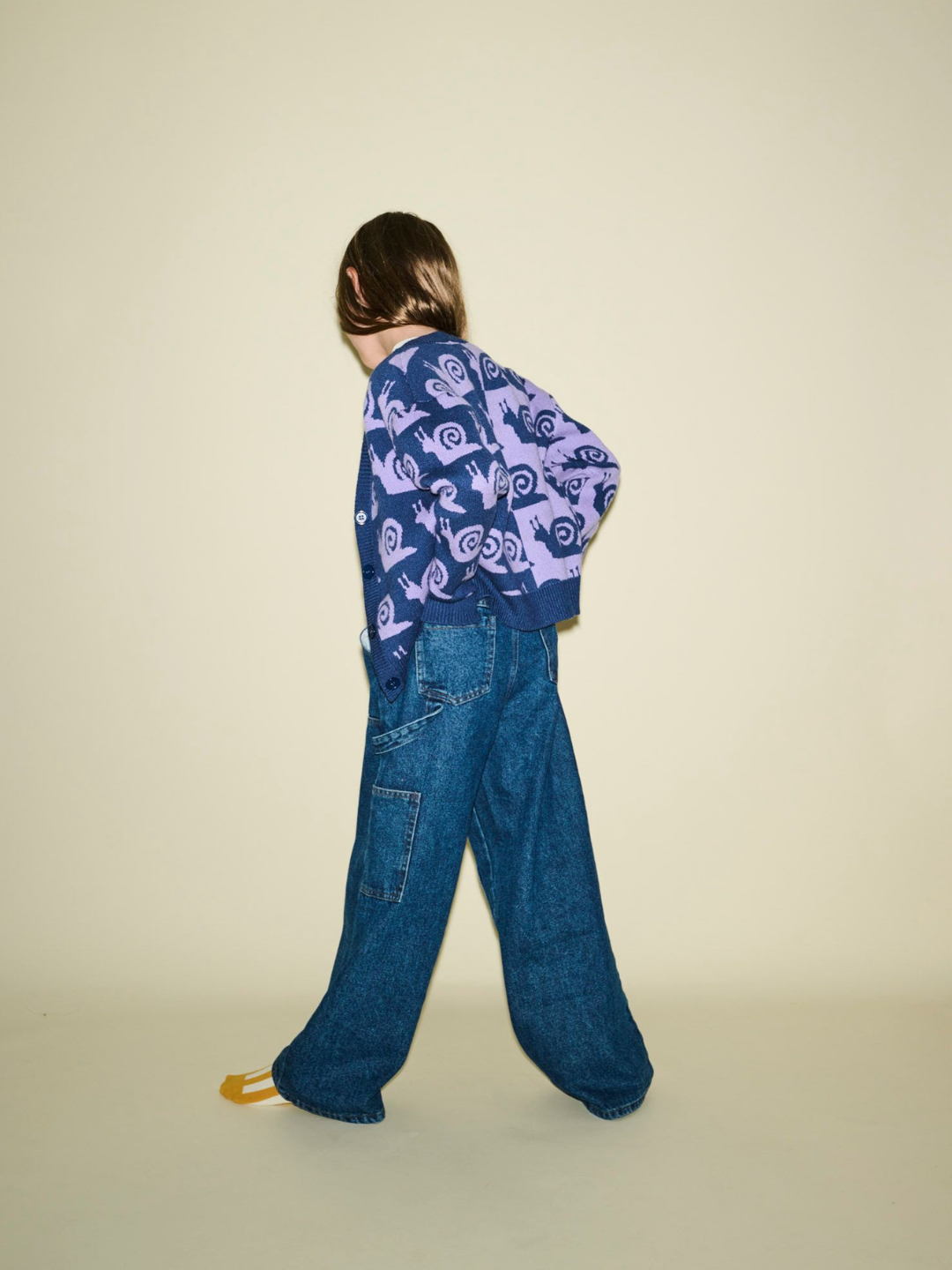 A person with long hair, wearing the ESCARGOT CARDIGAN and baggy jeans, stands against a plain beige background, turned slightly away from the camera.