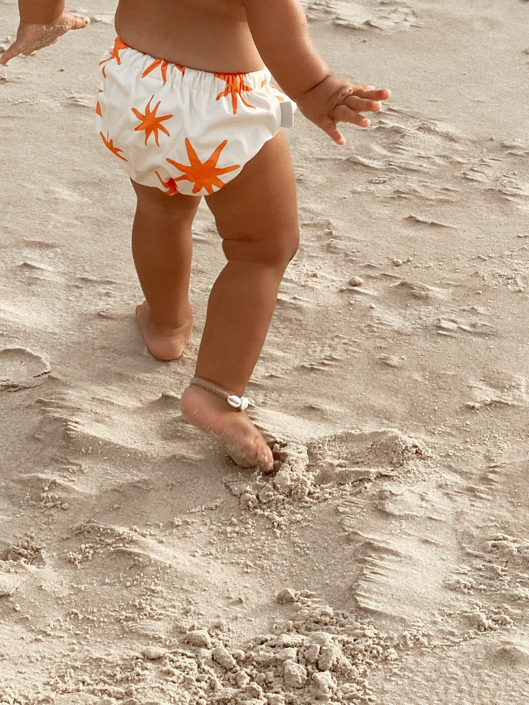 Capri | The back view of the swim diaper with an elastic waist with a tie and elastic leg holes on a child at the beach. The diaper is a cream color with orange stars shapes all over.