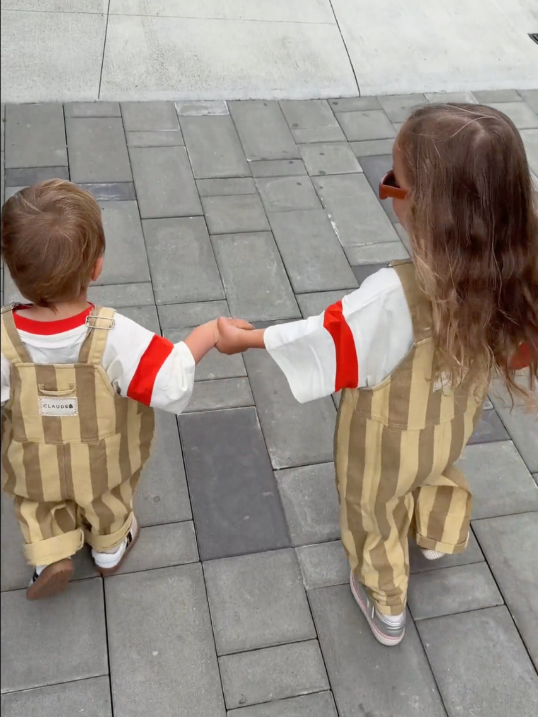 Two toddlers seen from behind holding hands while walking on a gray paved outdoor surface. Both are wearing matching beige and tan vertically striped overalls over white shirts with red sleeve stripes.
