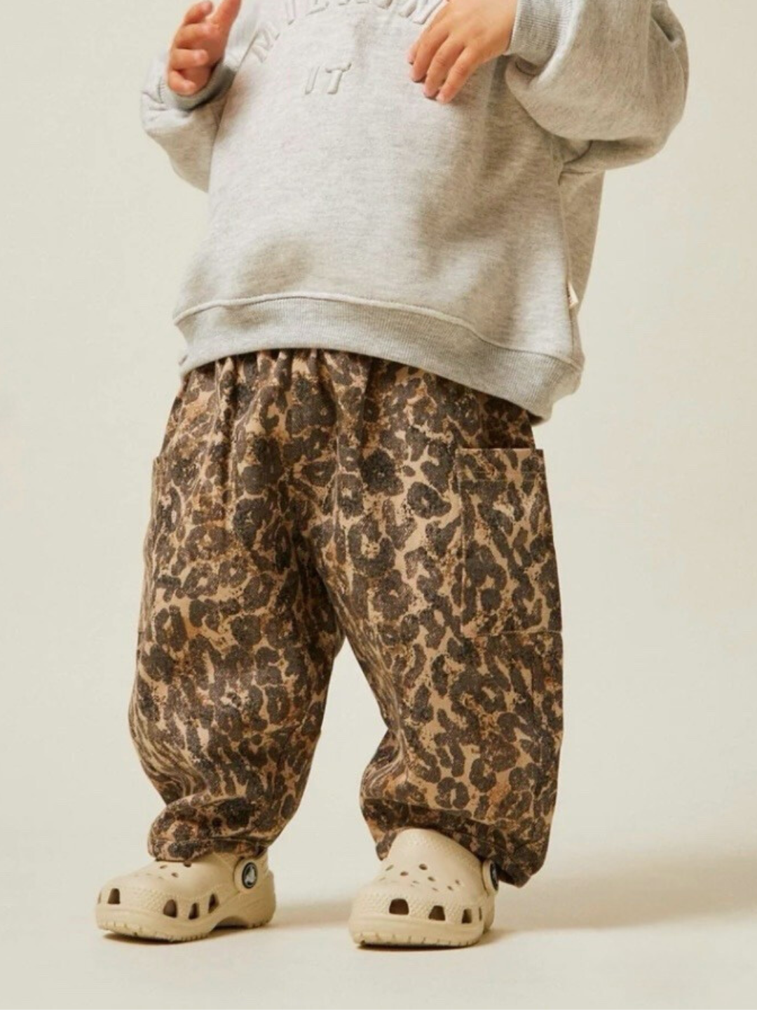 A young child stands against a neutral background, wearing a gray sweatshirt paired with EMERSON LEOPARD TROUSER made of organic cotton, and beige sandals.