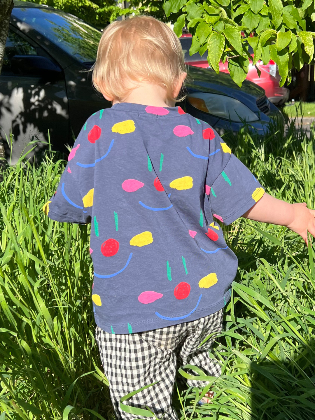 A toddler wearing the kids' Facepaint Tee in Navy, walking away from camera through long grass, and wearing black and white gingham pants.