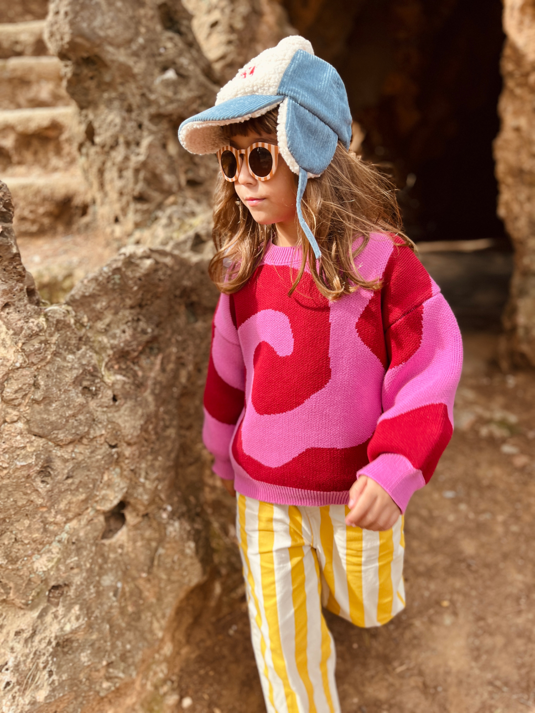 A girl wearing a kids crewneck sweater in bright pink, with a large single red swirl design that covers the entire sweater. She is is walking in front of a brown stone wall, and also wears yellow-and-white vertical striped pants, a blue cap with ear flats, and rust and white striped sunglasses.