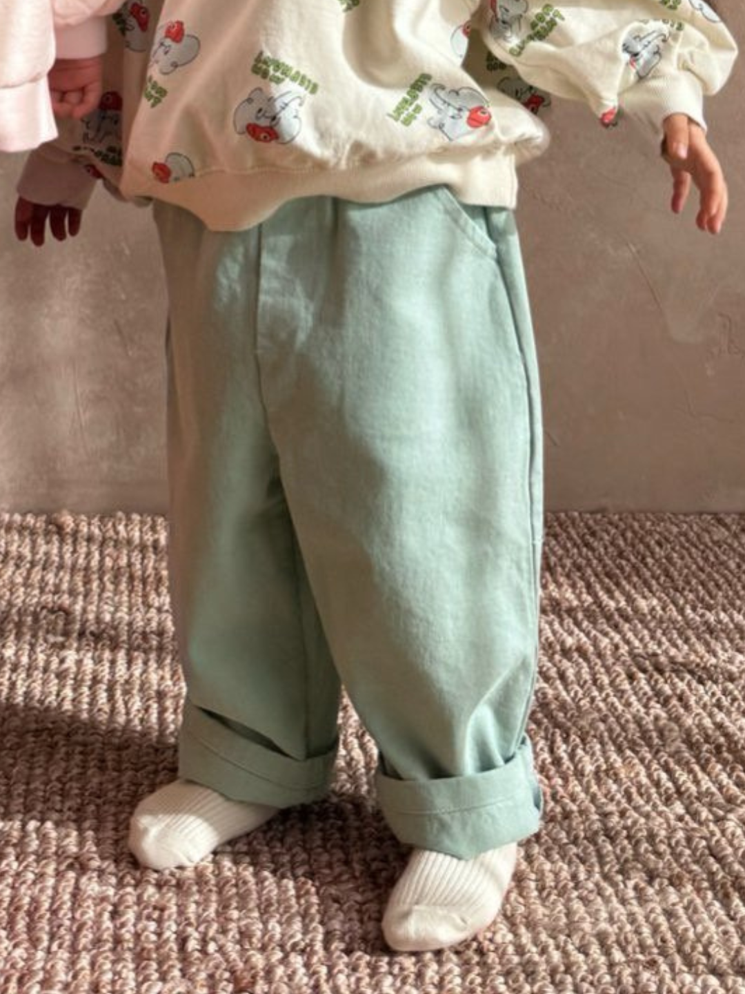 Mint | A child stands on a textured carpet, stylishly wearing Tomboy Pants in light green and a cream printed sweater, capturing the flair of a Korean kids brand.