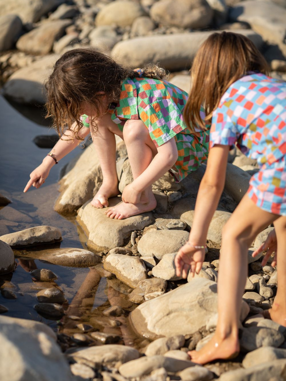 Two children by the water wearing kids' shirt and shorts sets in two different colorways: one with gold, orange, pink and purple squares on a green background; one with red, orange, pink, lilac and green squares on a blue background