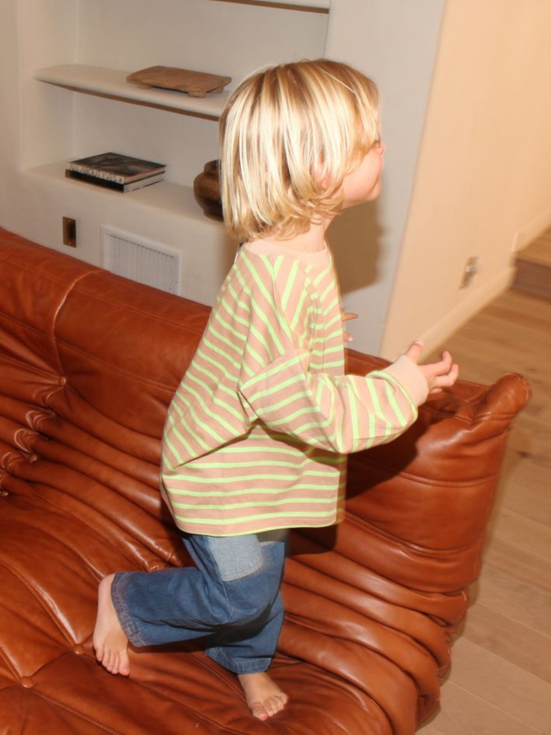 A blond child wears the BAILEY STRIPE LONGSLEEVE tee in beige with green stripes—with blue jeans, standing on a brown couch.