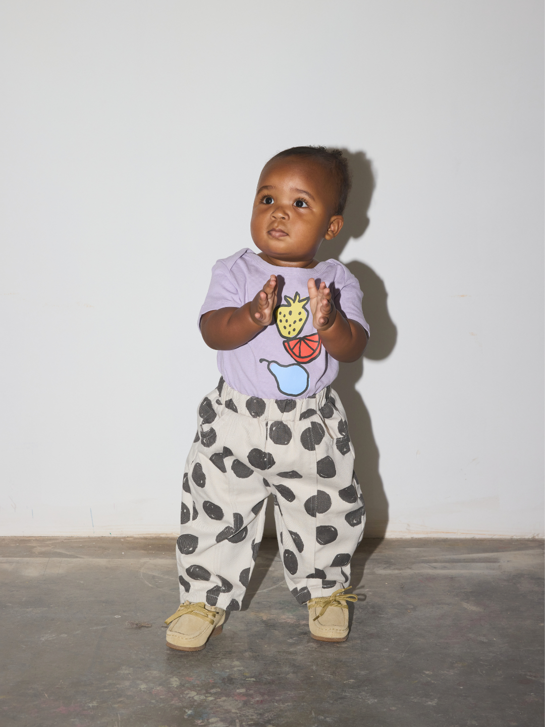 A toddler wears the Fruit Stack baby tee, featuring a strawberry, lemon slice and a pear, worn with polka dot jeans, standing on a grey floor in front of a white wall.