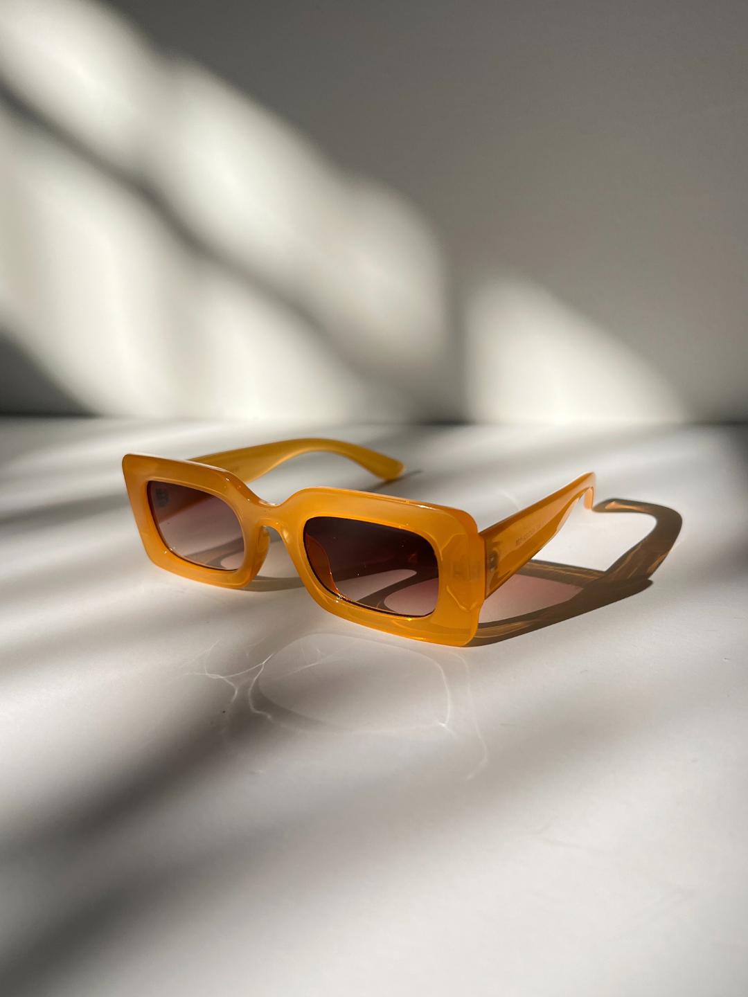 Kids rectangle sunglasses in orange, on a grey background with areas of sunlight, creating shadows of the sunglasses.