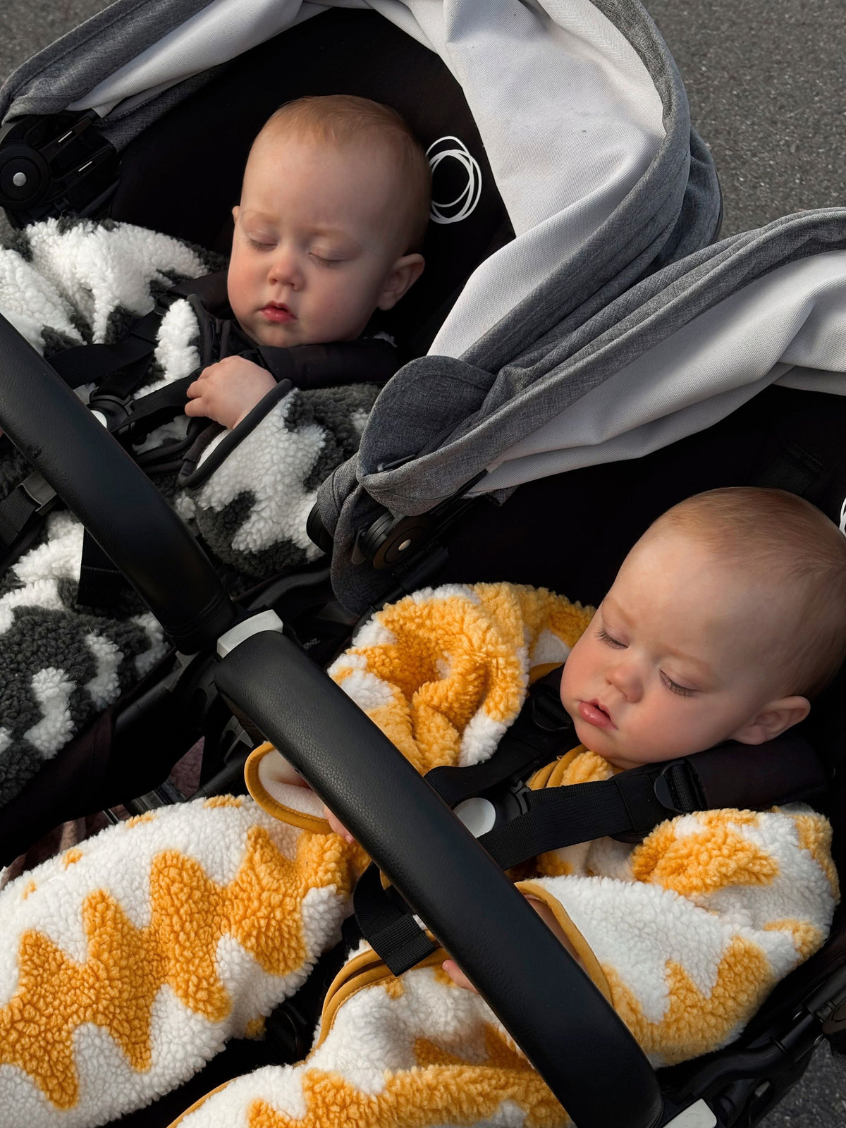 Two babies in a double stroller, wearing sherpa fleece winter suits, in a wave pattern, one in black and white, one in yellow