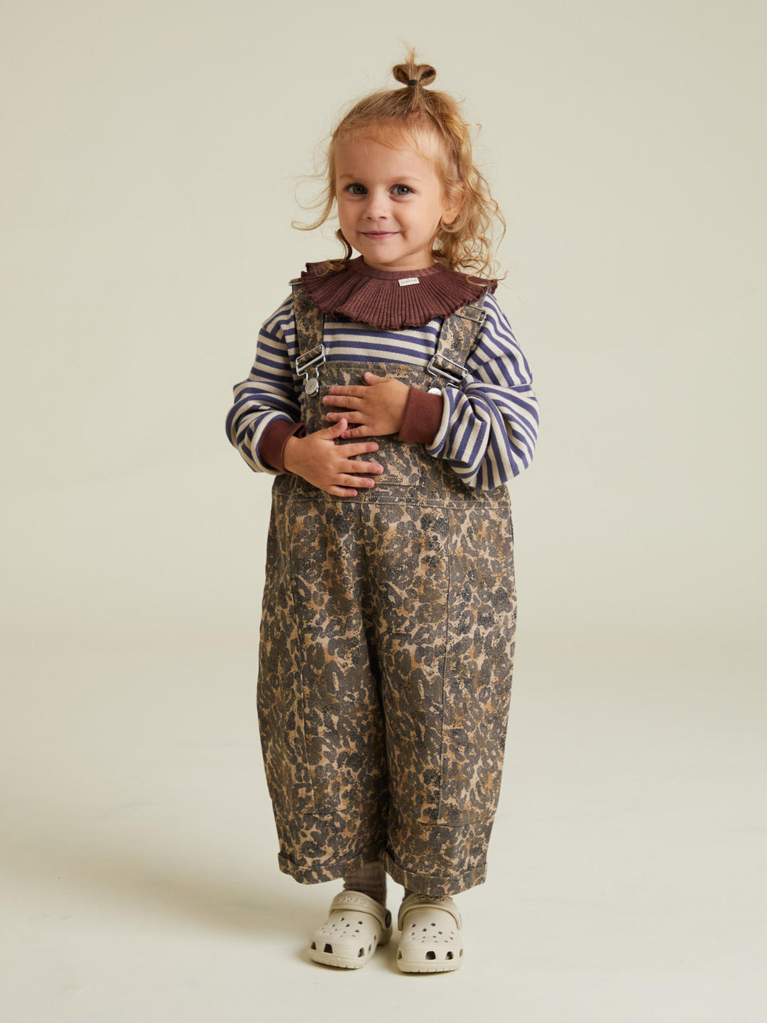 A young child with curly hair smiles indoors, wearing a striped shirt and the BLYTHE LEOPARD DUNGAREE with white slip-on shoes—no hidden shipping fees or extra costs included.
