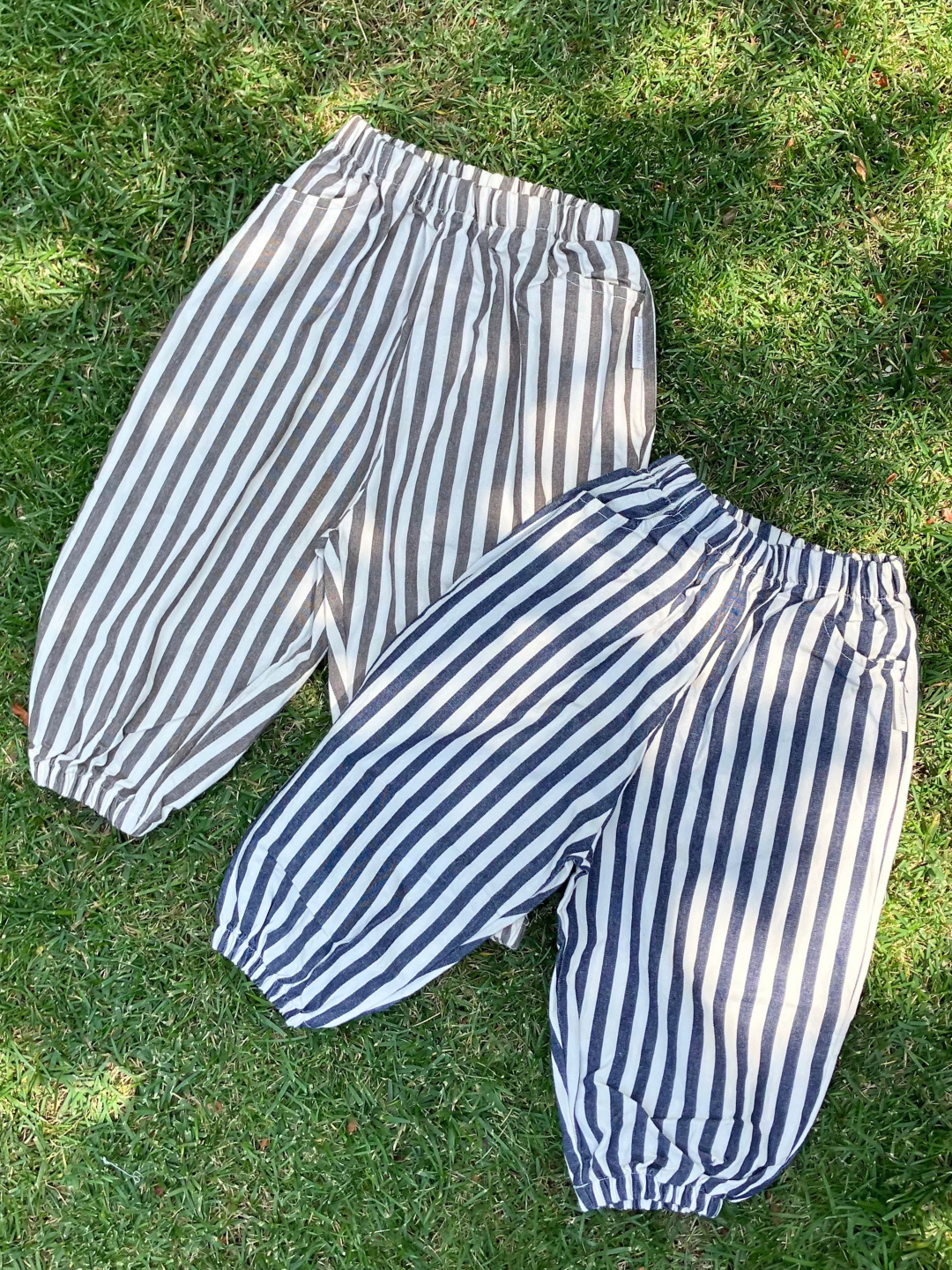 Two pairs of PIRATE PANTS from Minirobe, featuring striped cotton fabric with elastic waistbands and ankle cuffs, are laid side by side on grass. One pair showcases black and white stripes, while the other displays blue and white stripes, both with a stylish balloon leg design.