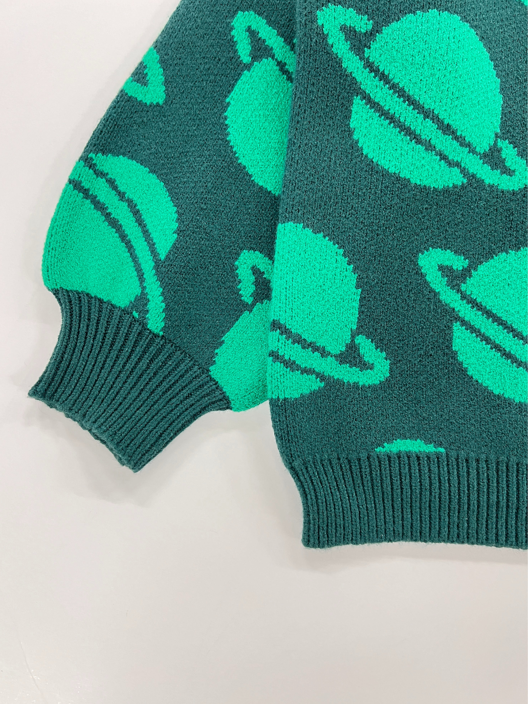 Green | A close-up photo of the SPACE CAMP SWEATER, a dark green garment featuring bright green planet-like shapes. The image highlights one sleeve and part of the body, focusing on the detailed cuff and hem.