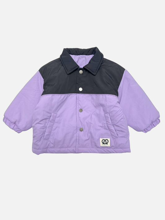 Image of A color-blocked kids puffer jacket with a black yoke and lavender-purple sleeves and body, featuring a snap front and a small pretzel logo patch on the lower left front.