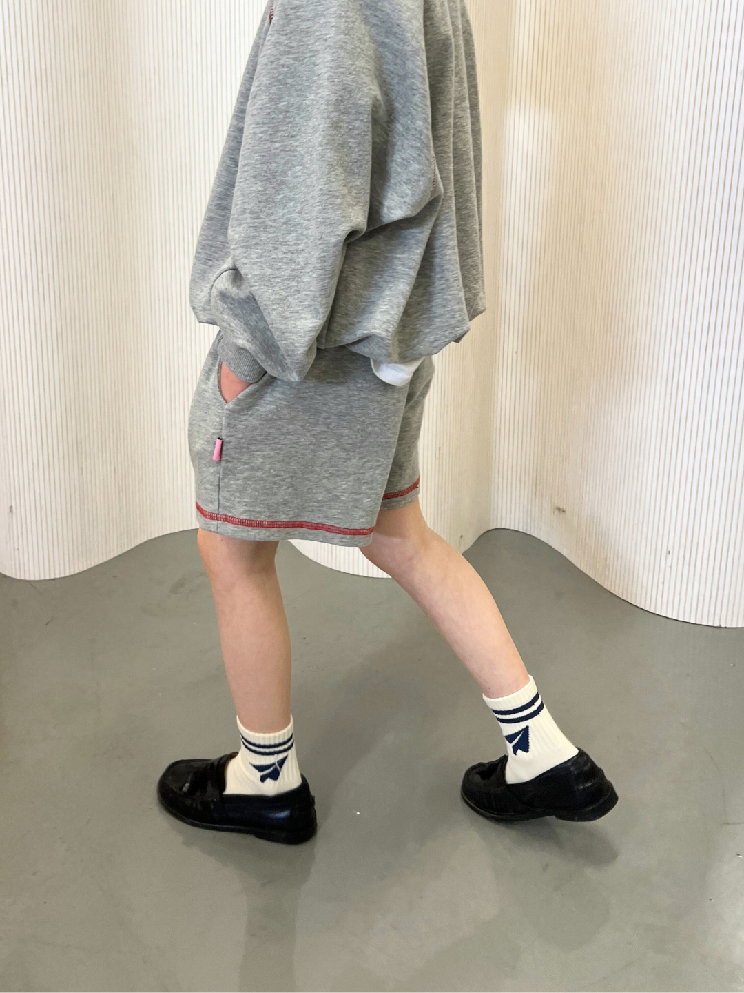 Grey | Dressed in a gray vintage-inspired outfit from a Korean kids' brand, the individual pairs VARSITY SWEAT SHORTS with a matching sweatshirt. Black shoes and white socks with blue stripes and bow accents complete the look over a white textured backdrop.