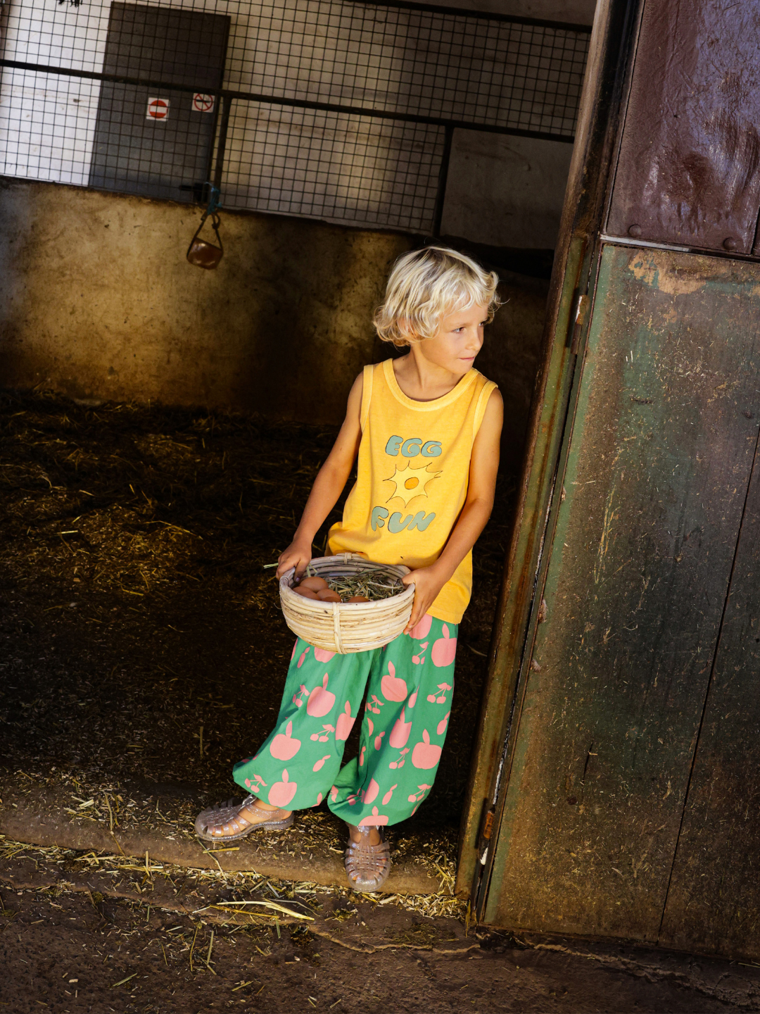 A child wearing vibrant FRUIT LOUNGE PANTS holds a basket, standing near the stable entrance.