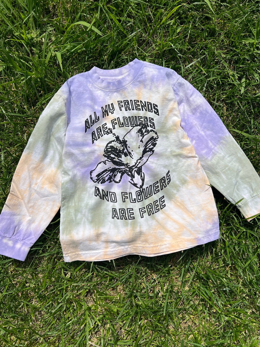 The ALL MY FRIENDS LONGSLEEVE, a pastel tie-dye longsleeve tee with a bold flower graphic and the phrase "All my friends are flowers and flowers are free," rests on grass in sunlight.