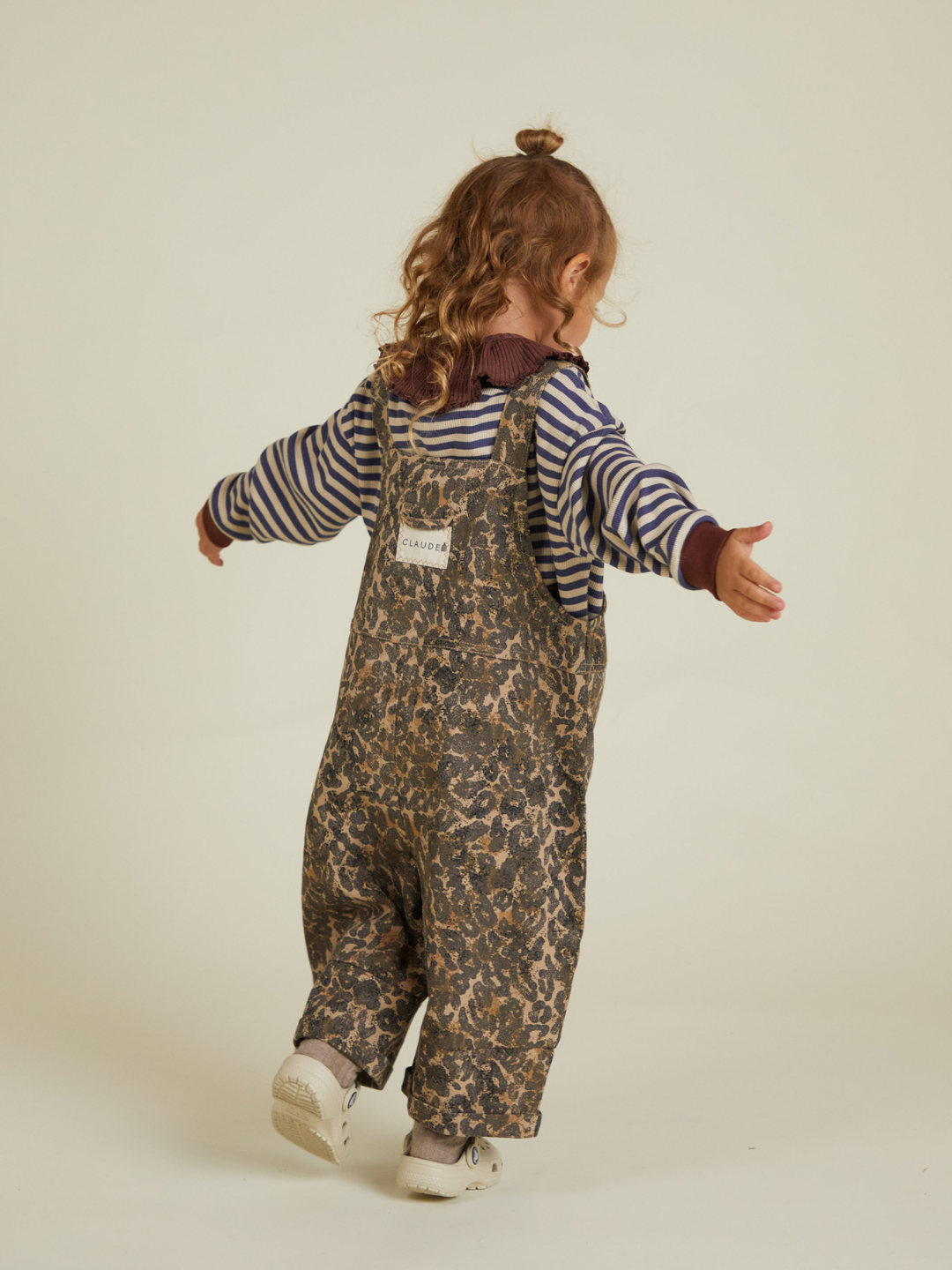 A toddler with curly hair wears the BLYTHE LEOPARD DUNGAREE over a striped long-sleeve shirt, standing with arms outstretched and facing away. Check shipping options and costs before purchasing.