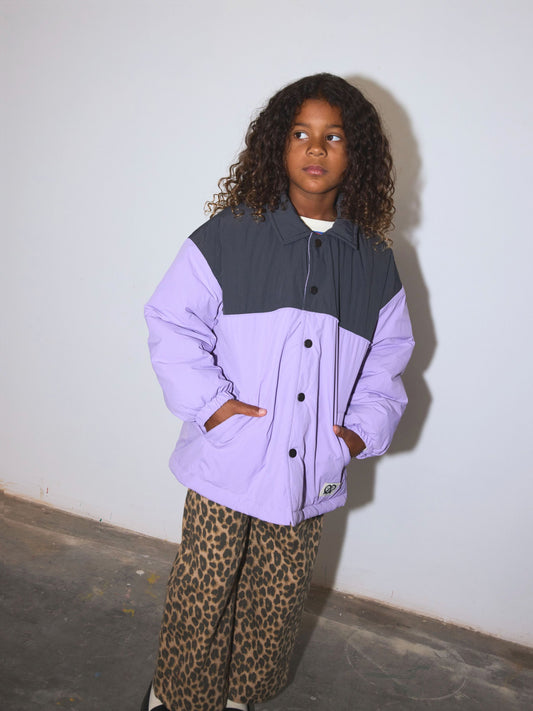 Second image of A color-blocked kids puffer jacket with a black yoke and lavender-purple sleeves and body, featuring a snap front and a small pretzel logo patch on the lower left front.