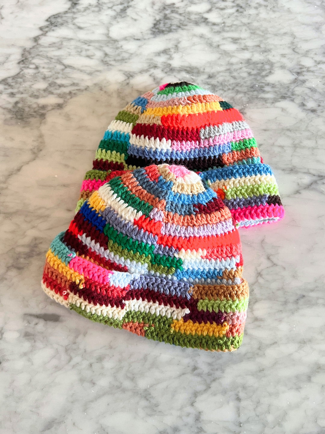 Two COLOR WHEEL BEANIE hats, featuring handmade multicolored stripes, are displayed on a gray marble surface.