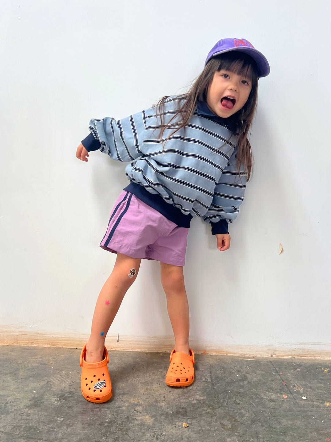 Lilac | A young child indoors wears SPRINTER SHORTS with a purple cap, striped sweatshirt, and orange Crocs, one arm bent and mouth open.