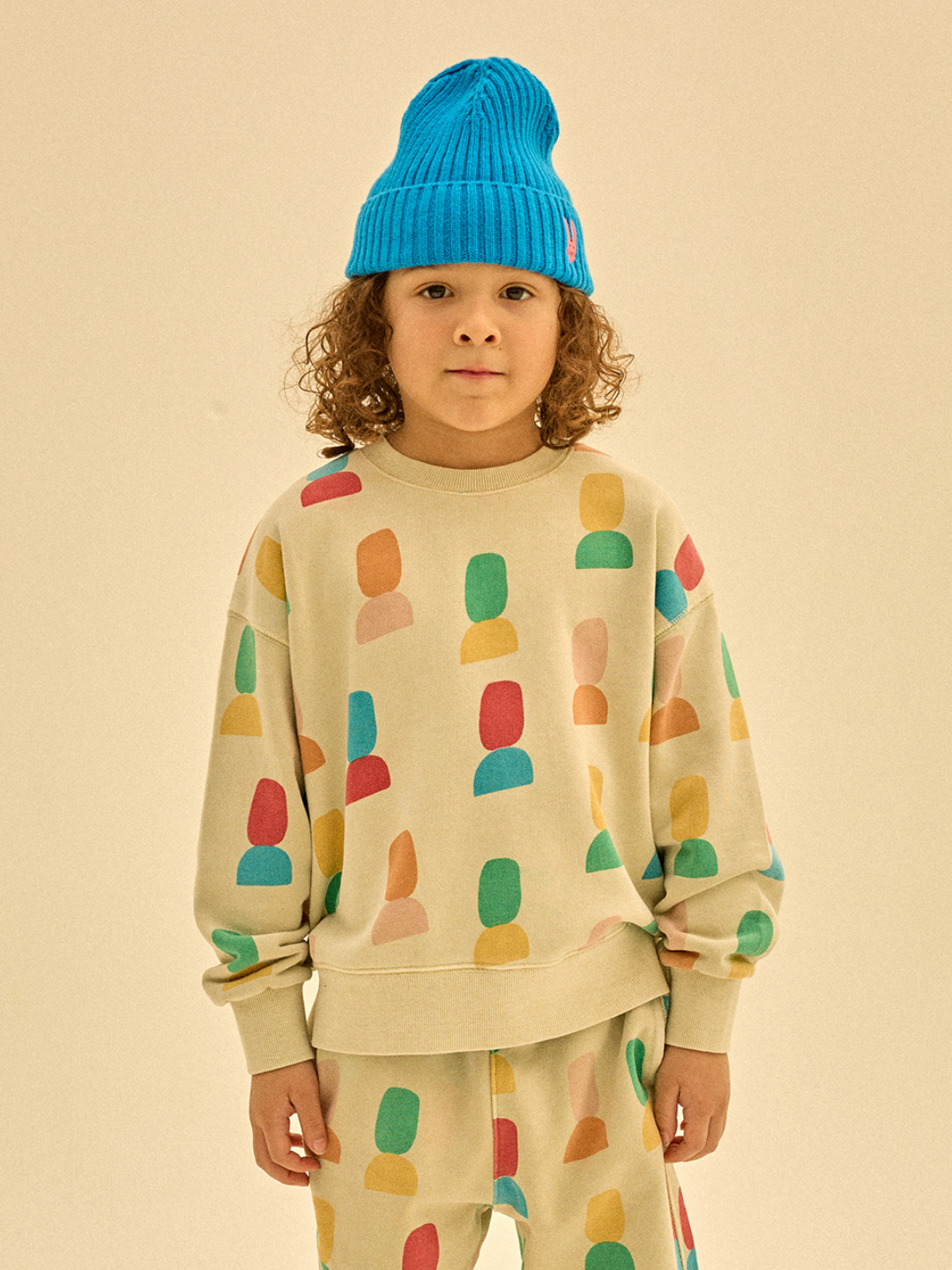 A child wears a vibrant, patterned outfit featuring the SHAPE PIGMENT SWEATSHIRT and matching pants, complemented by a bright blue knit hat. The plain beige background accentuates the meticulous merchandising.