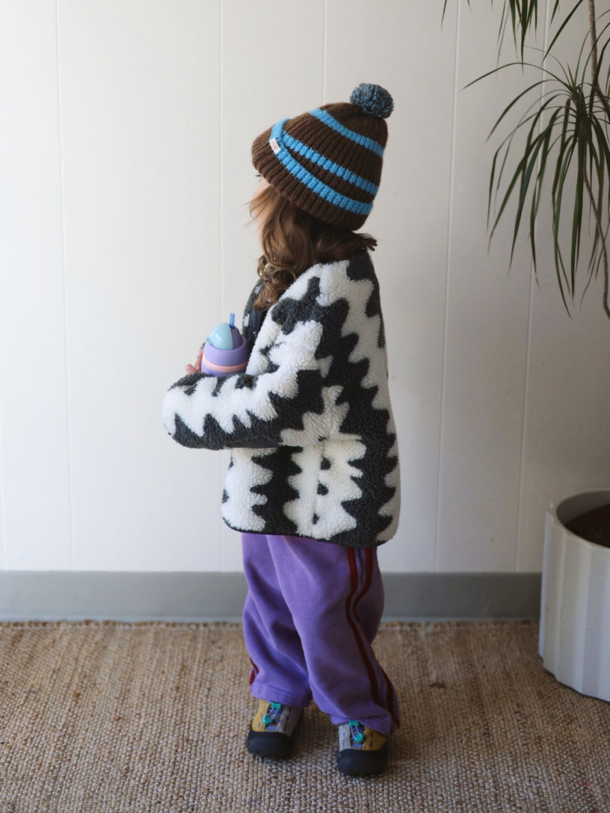 Child wearing a black and white wavy patterned jacket and blue hat against a white wall. She wears purple pants and stands on a jute rug