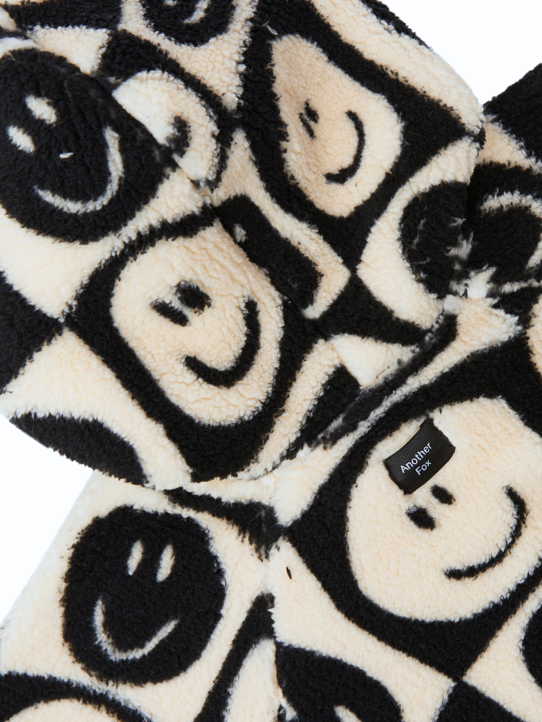 Close-up of a black and beige fluffy fabric with a pattern of smiley faces, perfect for baby clothing. A small label reading "Another Point" is sewn onto the fabric, making our adorable new arrival, the FACE CHECKER STROLLER SUIT, truly special.