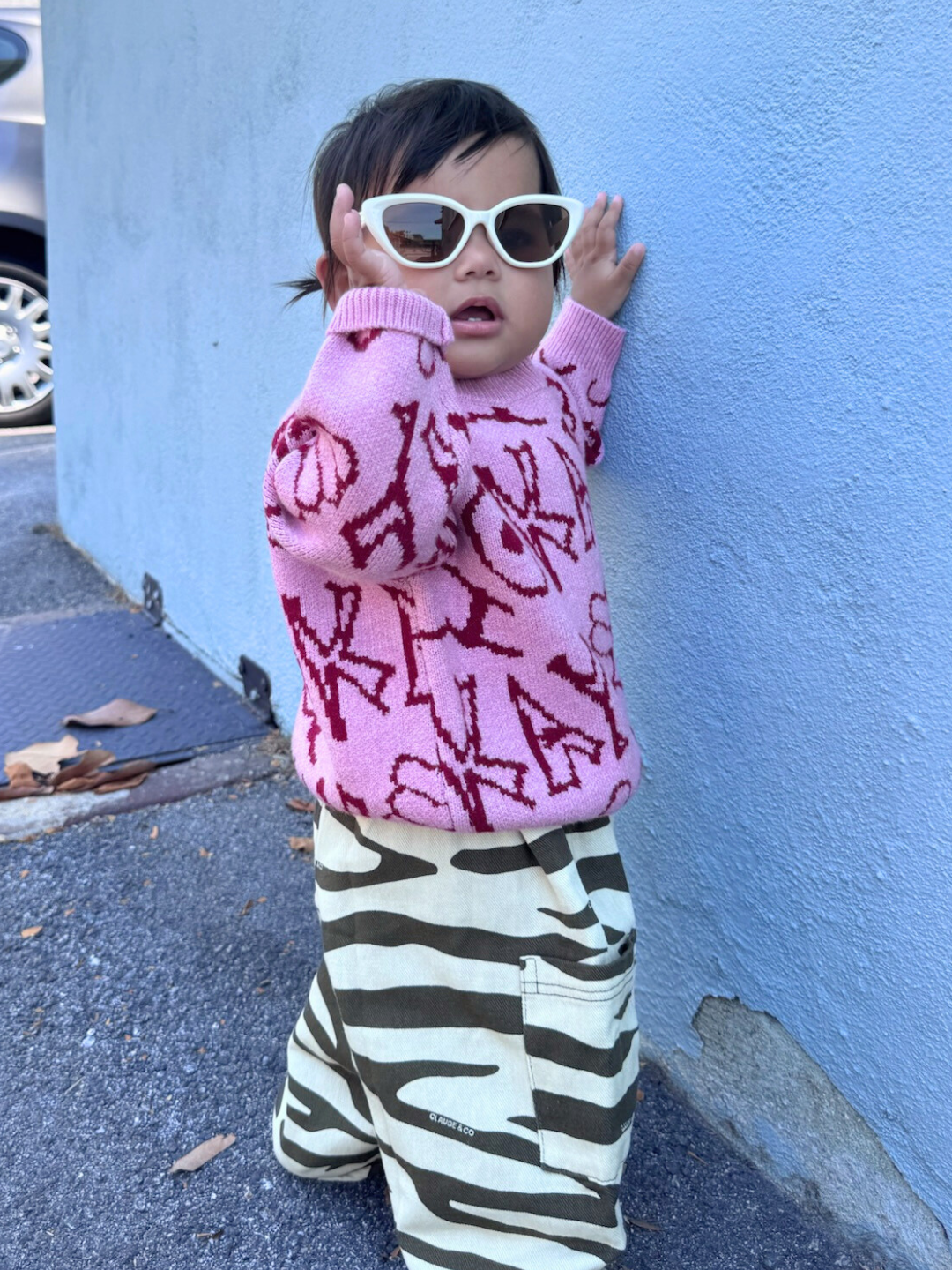 A young child wearing the OKAY SWEATER, zebra print pants, and white sunglasses stands outdoors against a blue wall.