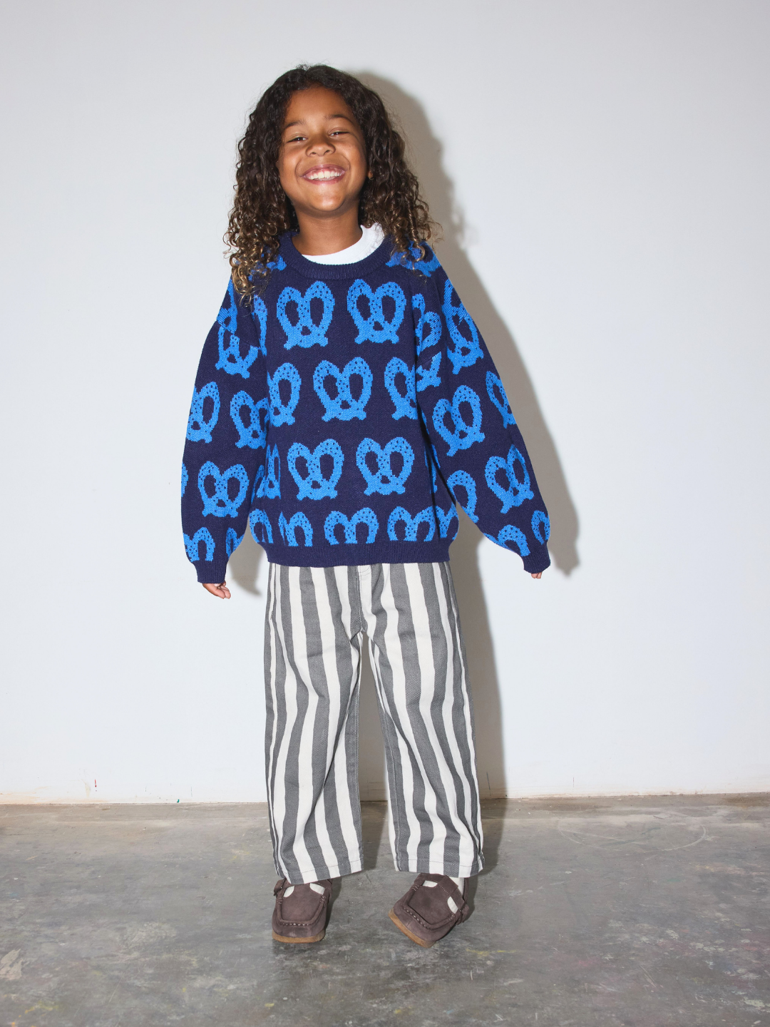 A smiling young girl with curly hair stands against a plain light gray wall wearing a navy blue long-sleeve sweater covered in a bright blue pretzel pattern, paired with gray and white vertically striped pants and brown shoes.