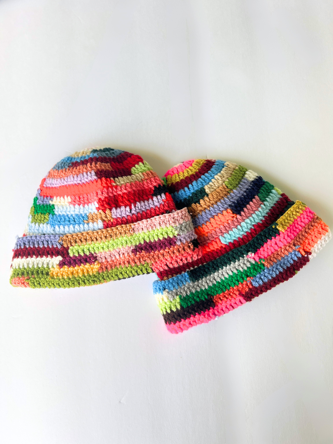 Two COLOR WHEEL BEANIE hats with vibrant patchwork designs and multicolored yarn are displayed on a white surface—handmade accessories that add a cheerful pop of color to any outfit.