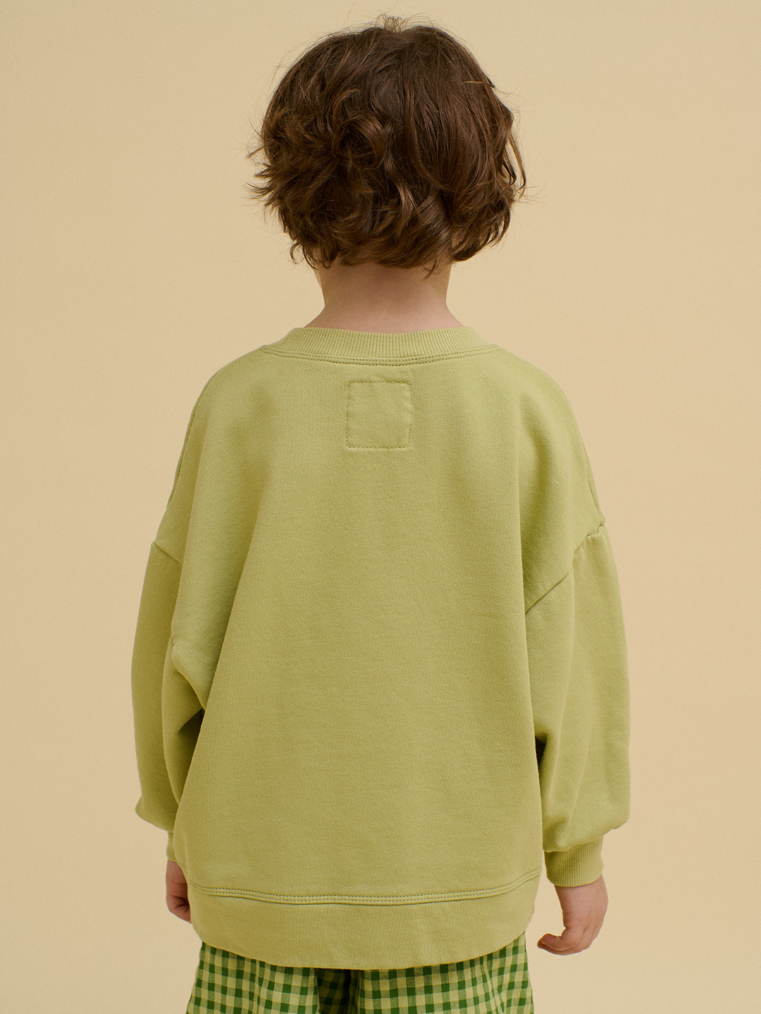 A child with medium-length brown hair stands with their back to the camera, wearing a STACKED POTATO SWEATSHIRT and green plaid pants against a beige background.