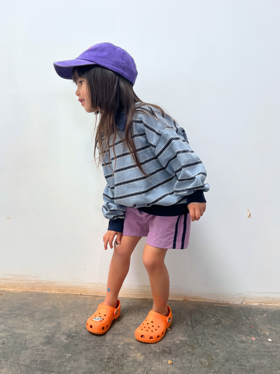 Lilac | A young child in a striped sweatshirt, SPRINTER SHORTS, purple cap, and orange clogs stands indoors against a plain wall, looking to the side.