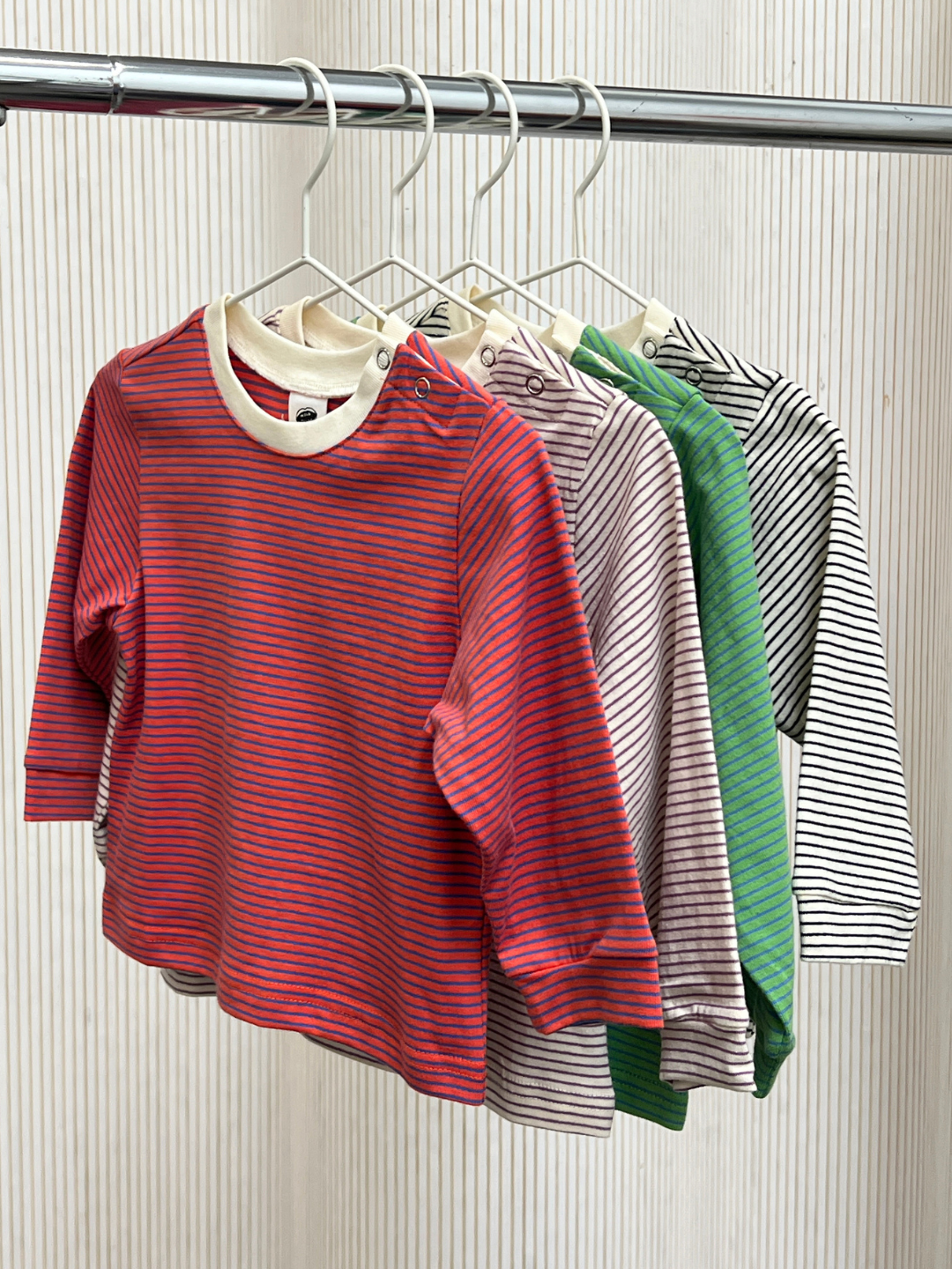 Four BABY STRIPE LONGSLEEVE shirts in red, beige, green, and white hang on white hangers from a metal rod against a light background. Made from soft cotton jersey, these basic baby and toddler tops are perfect for easy everyday dressing.