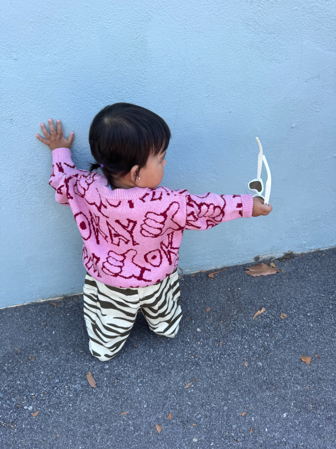 A toddler kneels on the ground facing a light blue wall, holding white sunglasses and wearing the OKAY SWEATER with red designs, paired with zebra-print pants.