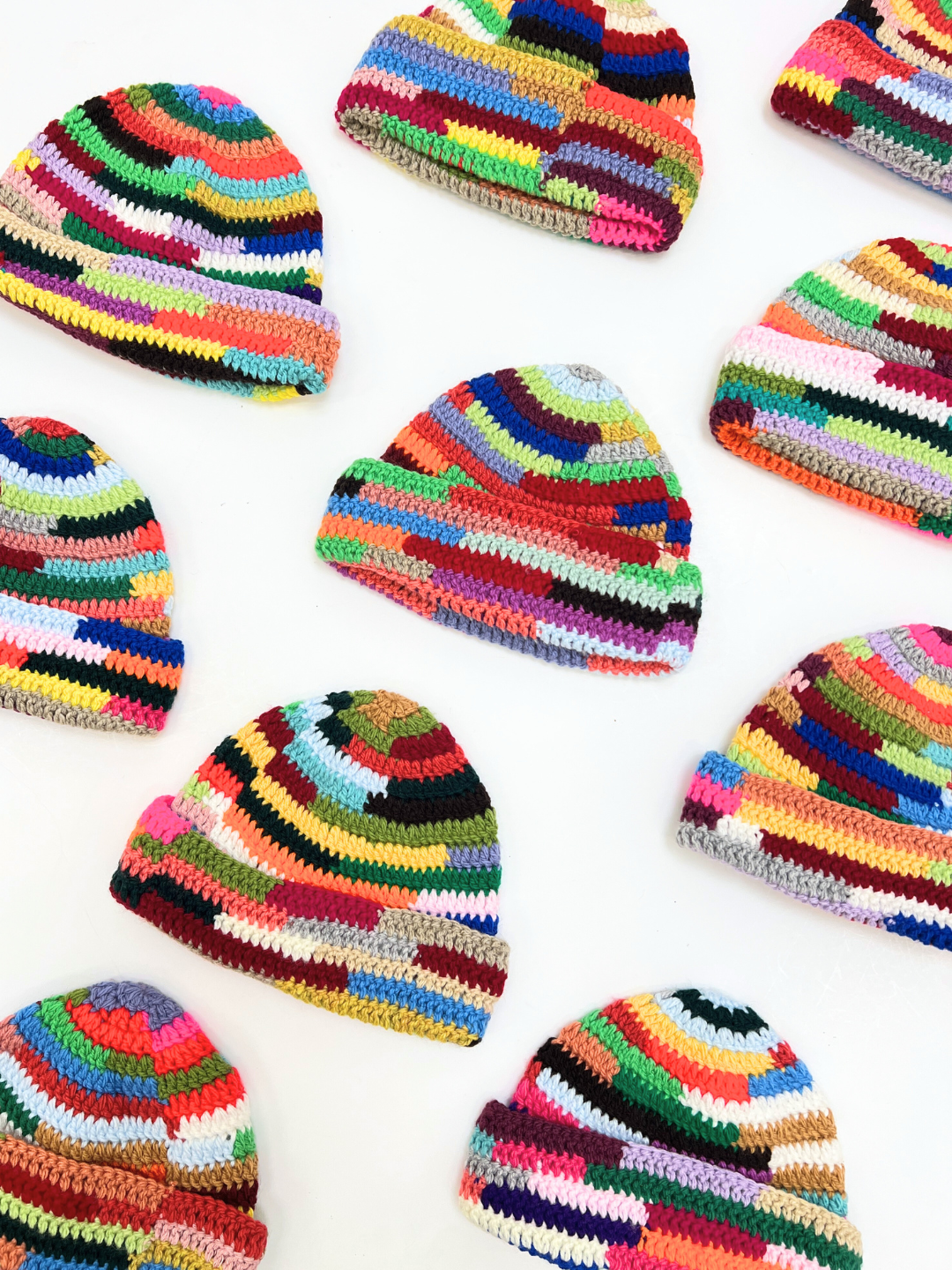 Nine COLOR WHEEL BEANIE hats, each handmade with vibrant stripes, are arranged in a loose grid on a white background, displaying their colorful styles.