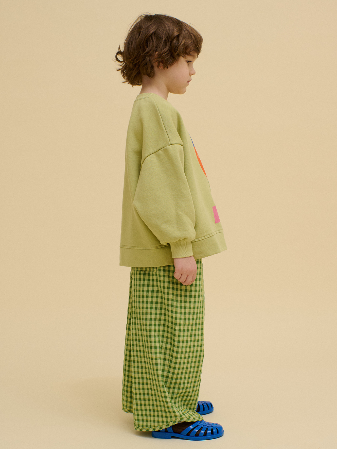 Child standing in profile wearing an oversized STACKED POTATO SWEATSHIRT, green checkered pants, and blue sandals against a plain beige background.