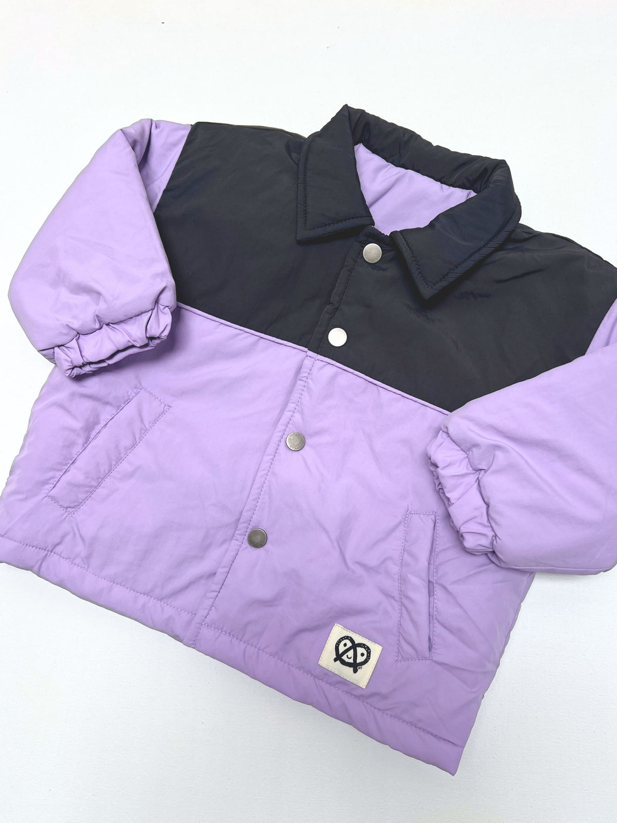 A kids lavender and dark gray colorblock puffer jacket, shown flat on a white background, featuring a small logo patch of a pretzel character