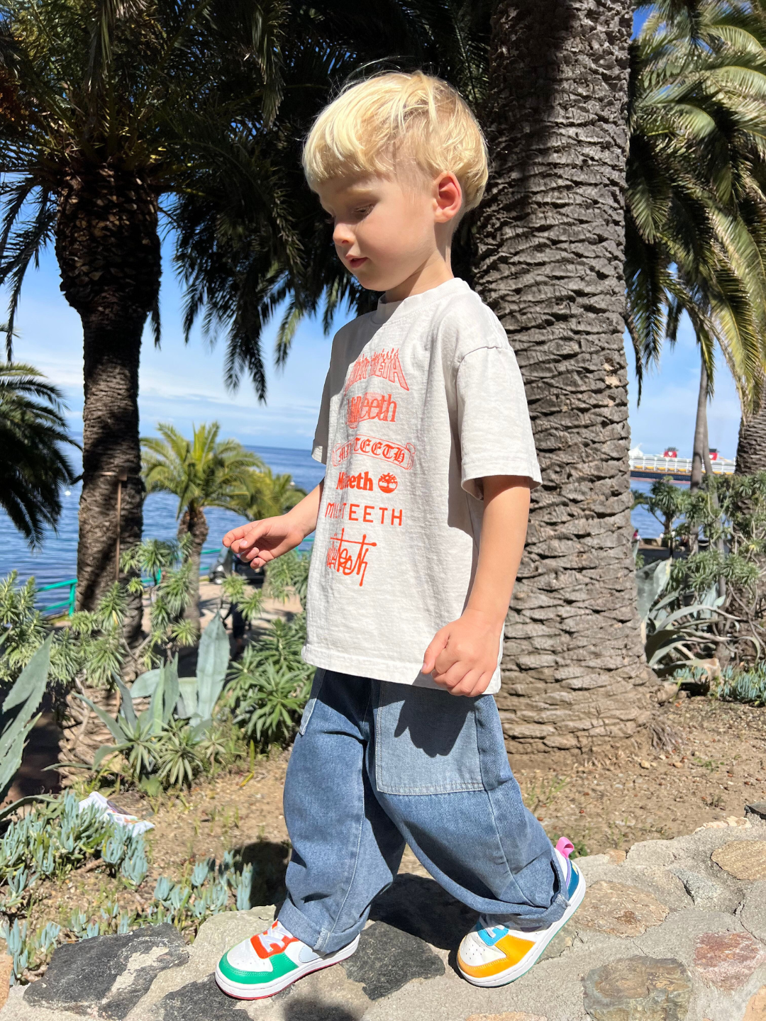 A child wearing the double trouble kids jeans in medium blue denim with lighter blue patch pockets. They have short blond hair, and are wearing a light grey tshirt with orange Milk teeth lettering, and white sneakers with colorful trim, walking on a stone wall with palm trees, the ocean and blue sky in the background.