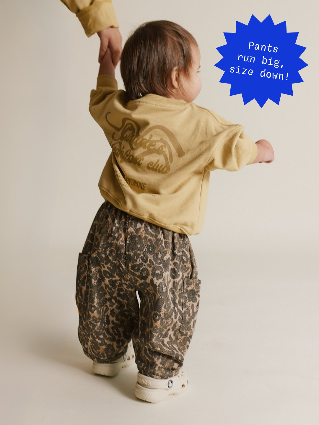 A toddler in a beige shirt and the cute EMERSON LEOPARD TROUSER walks while hand in hand with an adult. A blue sticker calls out that these pants run large, and to size down.