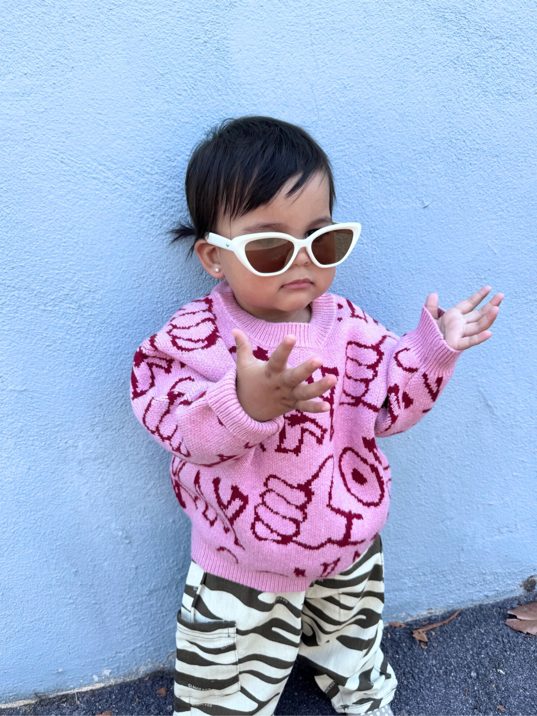 Pink | A toddler wearing white sunglasses, the OKAY SWEATER in pink with red patterns, and zebra-print pants stands in front of a light blue wall with arms spread.