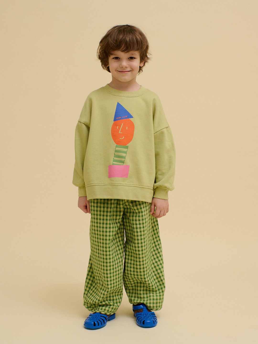 A child stands against a beige background, wearing an OXOX CLUB STACKED POTATO SWEATSHIRT in light green with a colorful geometric face, paired with green checkered pants and blue sandals.