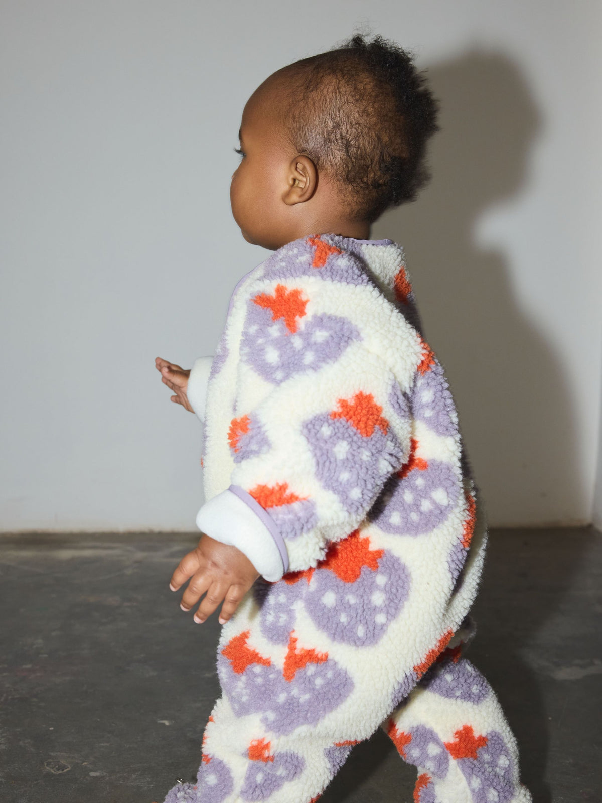 A baby wearing a cream, purple, and orange STRAWBERRY SHERPA FLEECE STROLLER SUIT with a strawberry print.