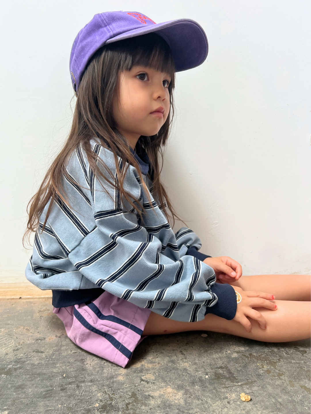 A young child with long hair wears SPRINTER SHORTS, a purple cap, and a striped sweatshirt while sitting on the floor against a plain white wall.