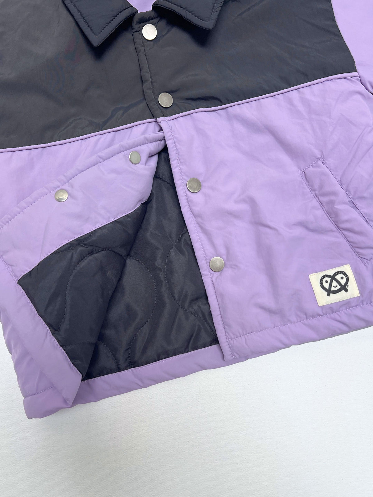 A close-up of a color-blocked puffer jacket with a lavender purple lower half and a dark gray upper half, featuring silver snap buttons and a small white logo patch on the front pocket.