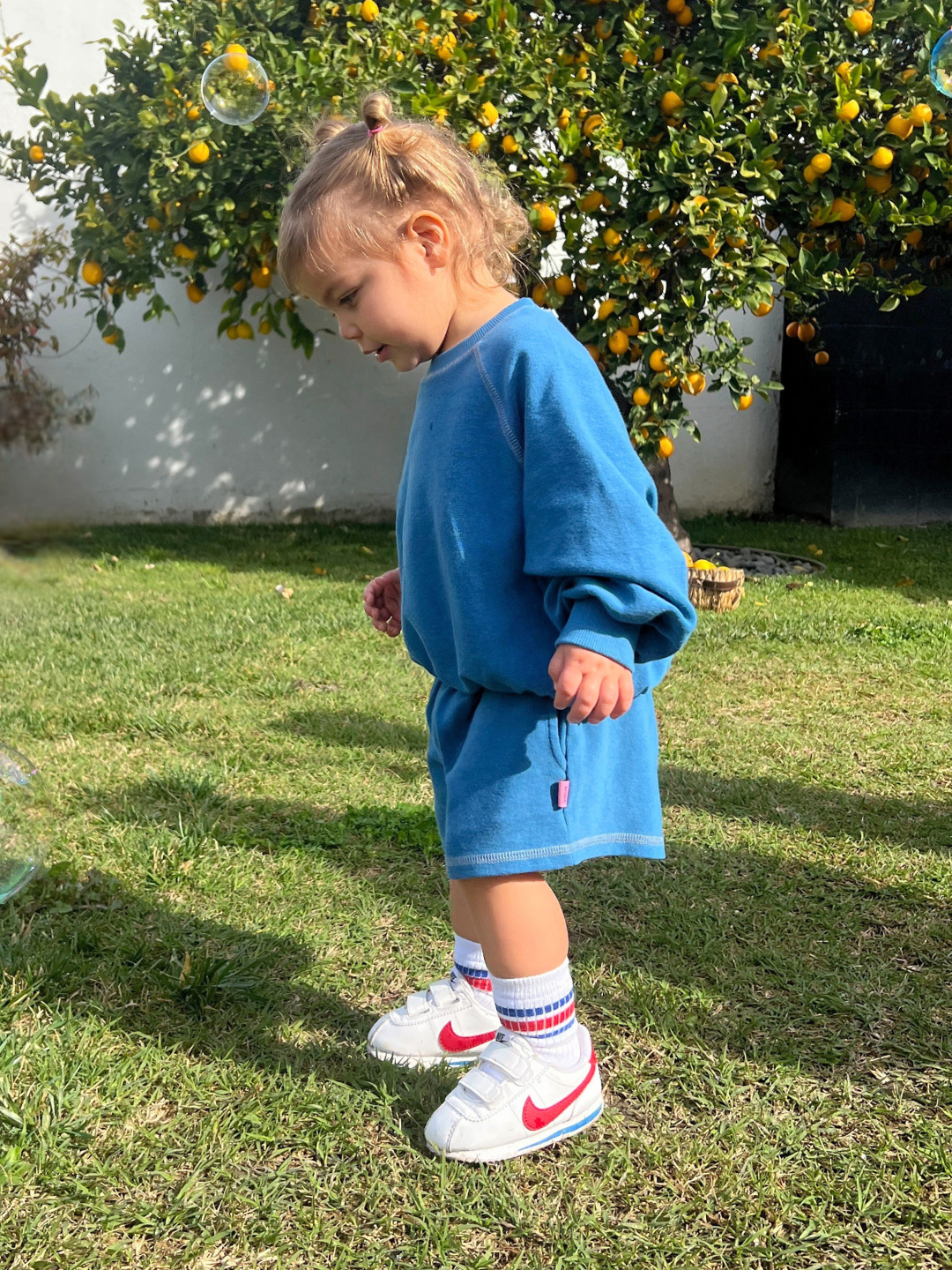 Blue | A toddler in a blue outfit and white sneakers stands on grass gazing at bubbles, with a citrus tree in the background, wearing brushed cotton VARSITY SWEAT SHORTS from a Korean kids brand.