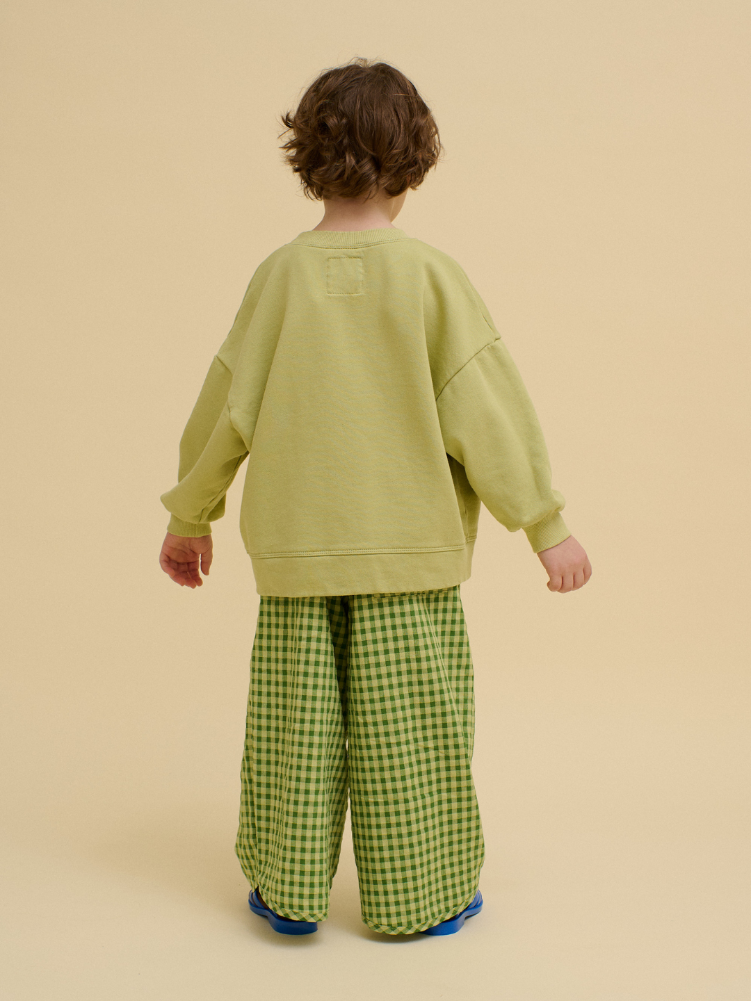 A child stands facing away, wearing a light green STACKED POTATO SWEATSHIRT and green checkered pants, set against a plain beige background.