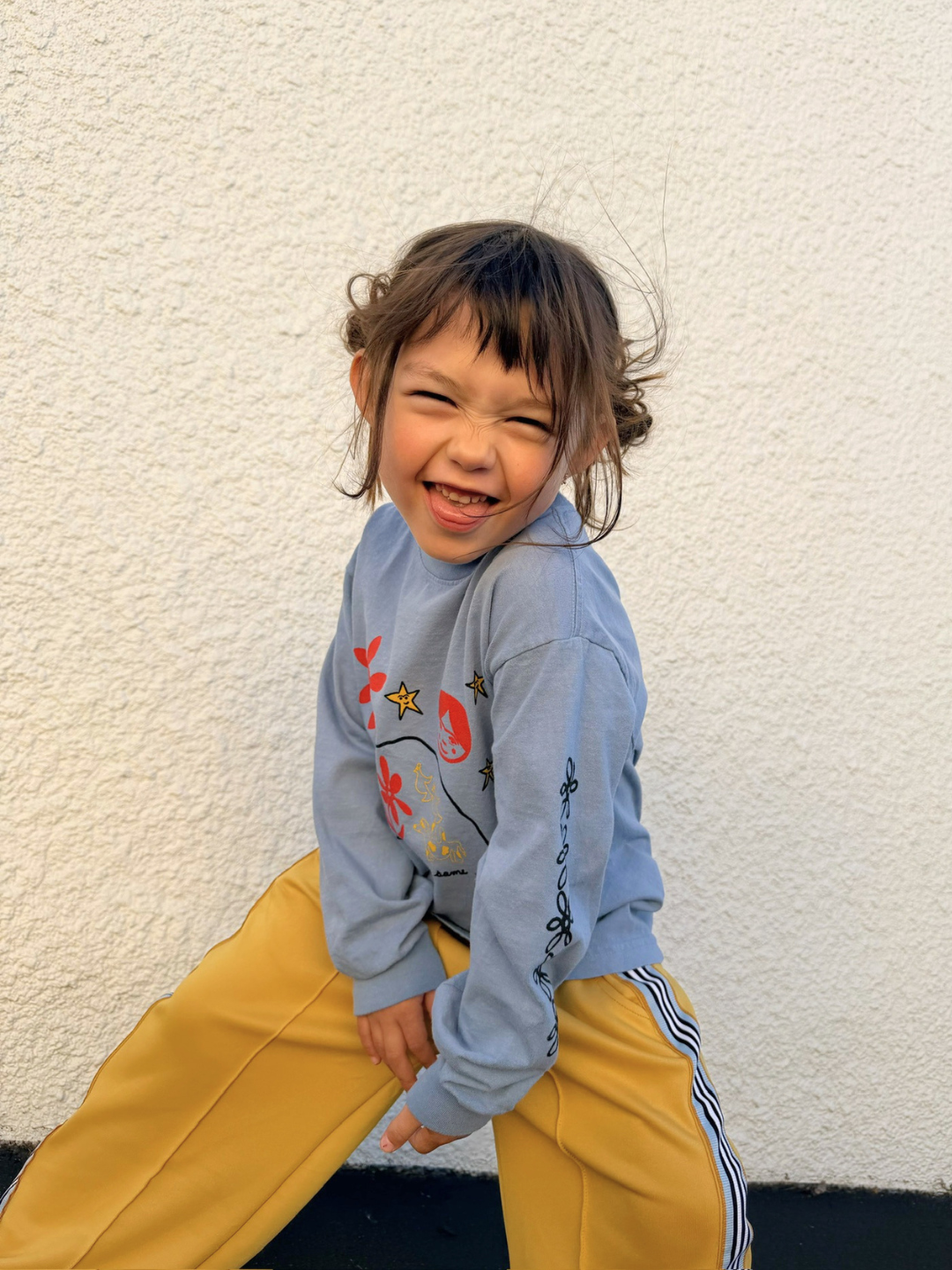 Blue | Child wearing a blue sweater with colorful designs and yellow pants against a white wall.