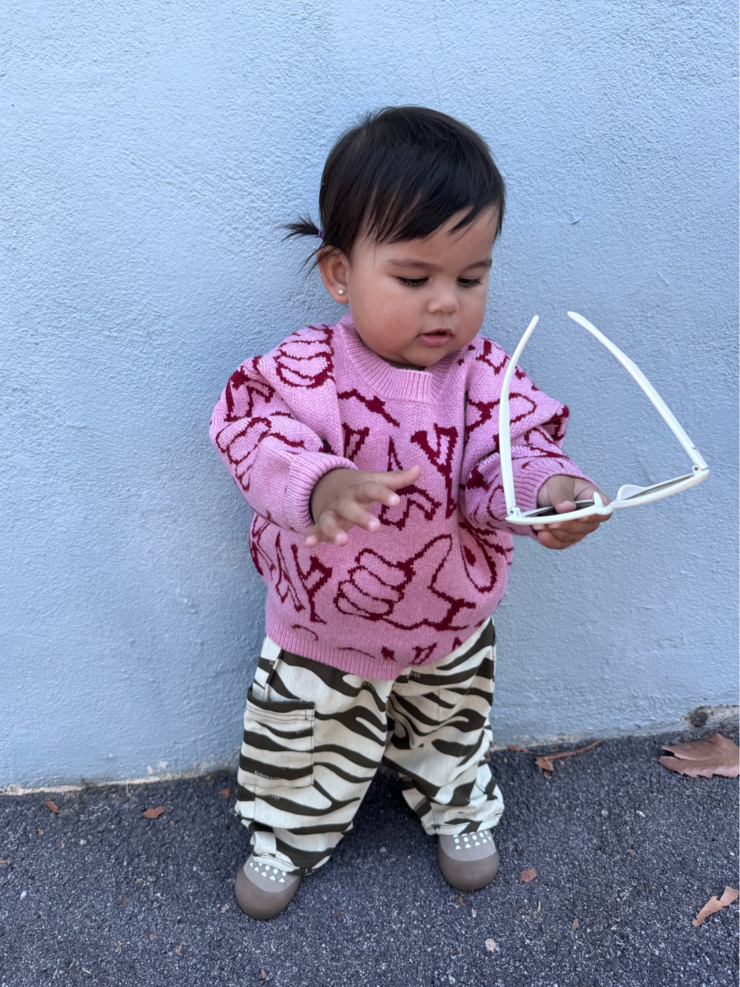 A toddler stands against a light blue wall, wearing the OKAY SWEATER—a pink patterned top—paired with zebra-print pants and holding white sunglasses.